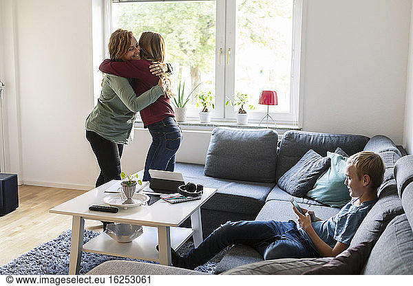 Smiling mother embracing daughter while son using phone at home