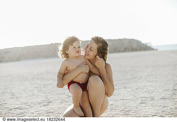 Smiling mother embracing son kneeling at beach enjoying vacation