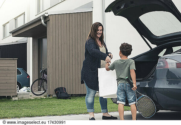 Smiling mother giving bag to son standing by electric car at front yard