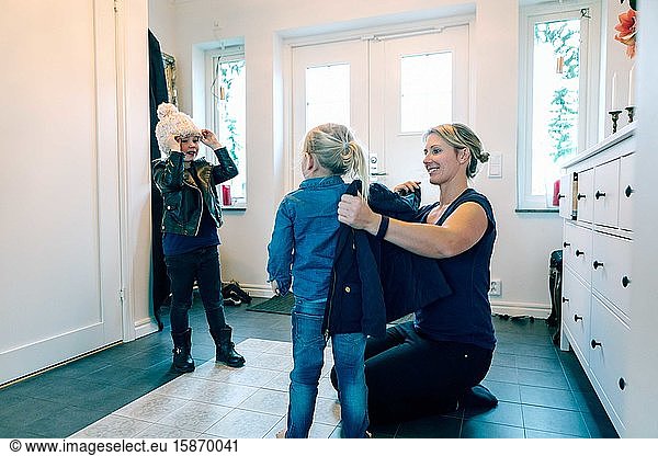 Smiling mother helping daughter with jacket while kneeling at home