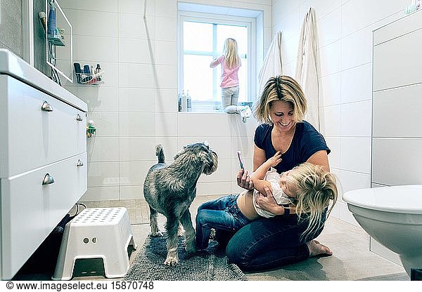 Smiling mother holding daughter with toothbrush by dog in bathroom