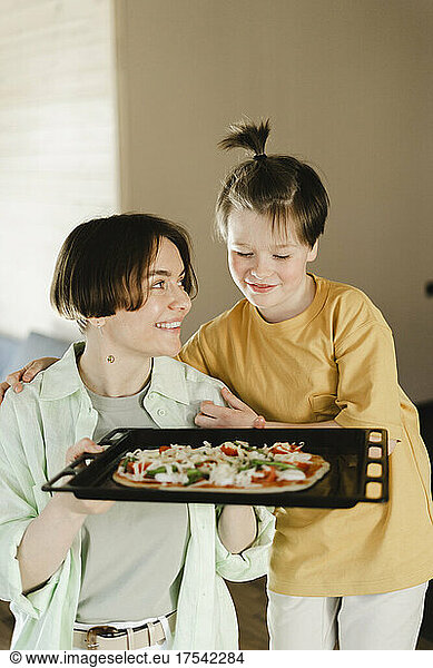 Smiling mother holding pizza dough in tray looking at son with ponytail