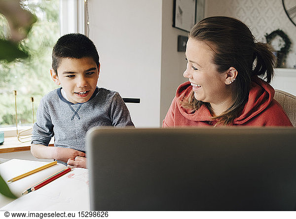 Smiling mother looking at autistic son watching video over laptop while sitting in living room