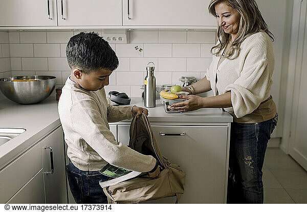 Smiling mother looking at son putting book inside bag in kitchen