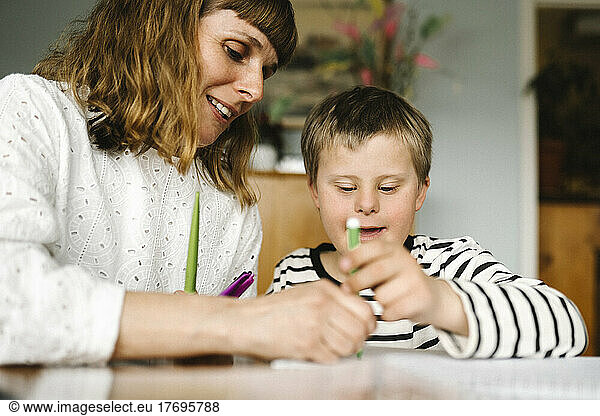 Smiling mother teaching son with disability to draw in book at home