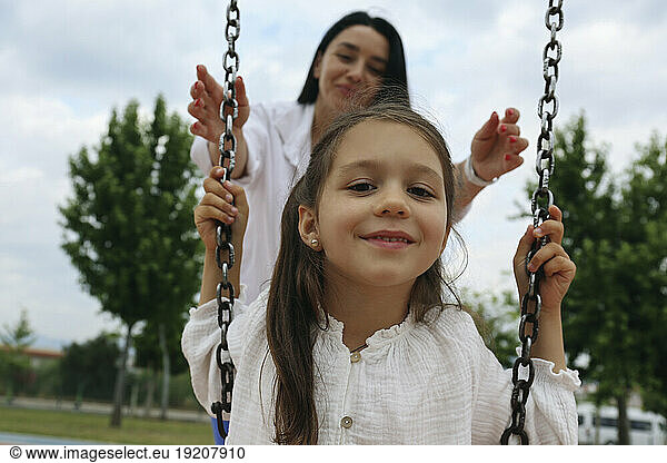Smiling mother with daughter sitting on swing in park