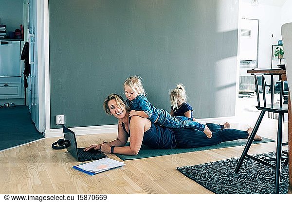 Smiling mother with laptop looking at daughter lying on her back while girl sitting in living room at home