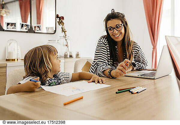 Smiling mother with laptop looking at daughter painting on table in dining table