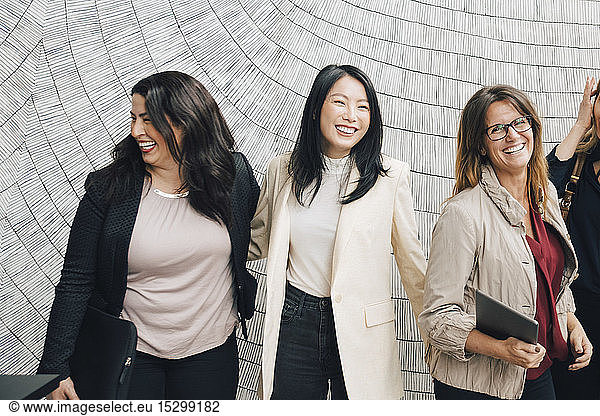 Smiling multi-ethnic female business professionals standing against wall at office
