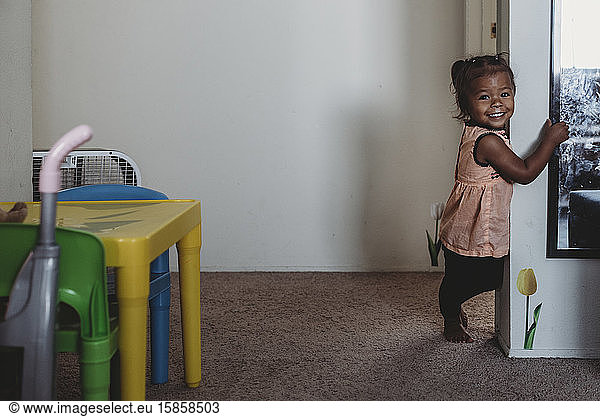 Smiling multiracial 2 yr old girl standing on carpet next to wall