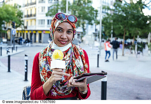 Smiling Muslim female journalist holding digital tablet while reporting in city during pandemic