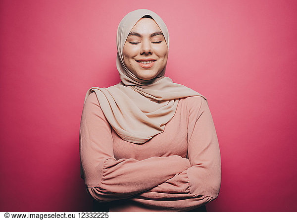 Smiling Muslim woman with eyes closed standing against pink background