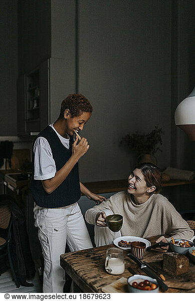 Smiling non-binary person brushing teeth by friend having breakfast at home
