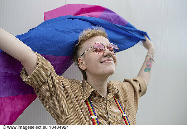 Smiling non-binary person holding pride flag in front of wall