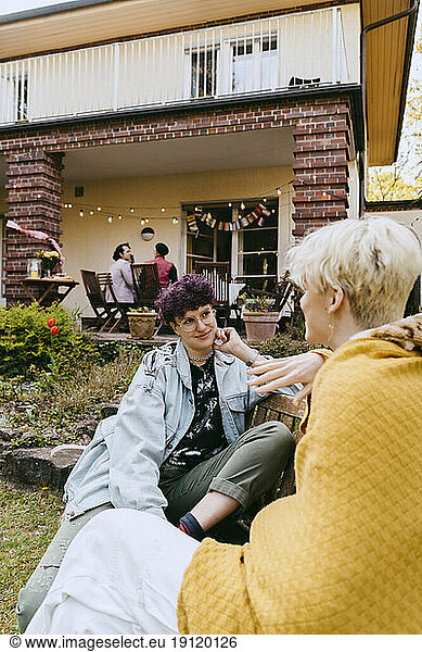 Smiling non-binary person talking to gay friend while sitting on bench outside house