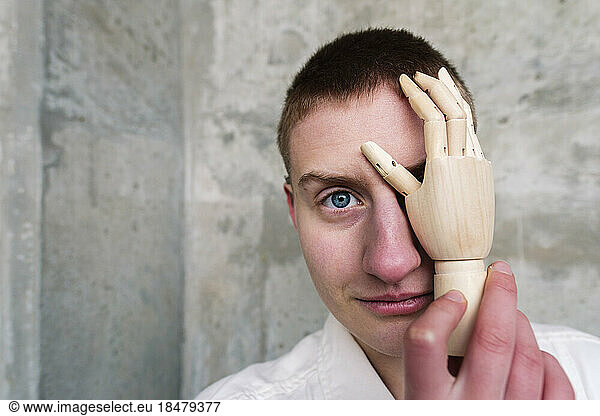 Smiling non-binary person touching face with robotic hand in front of wall