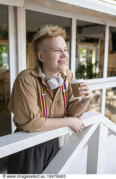 Smiling non-binary person with disposable coffee cup at railing