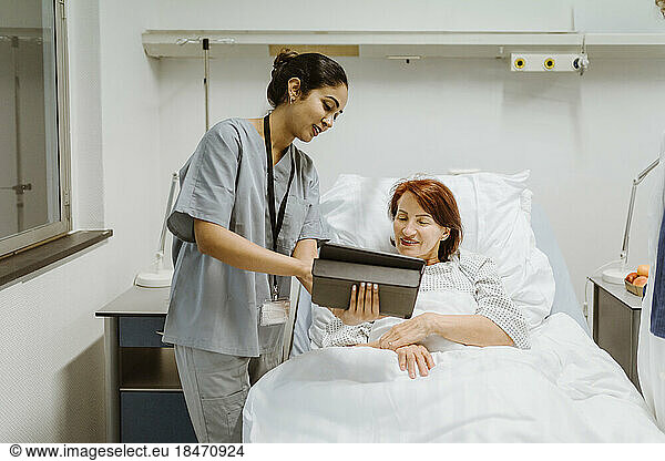 Smiling nurse showing digital tablet to senior female patient lying on bed in hospital