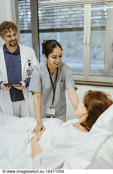 Smiling nurse talking to senior female patient lying on bed by doctor in hospital