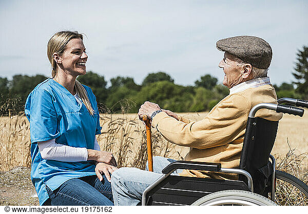 Smiling nurse talking with elderly man sitting in wheelchair on sunny day