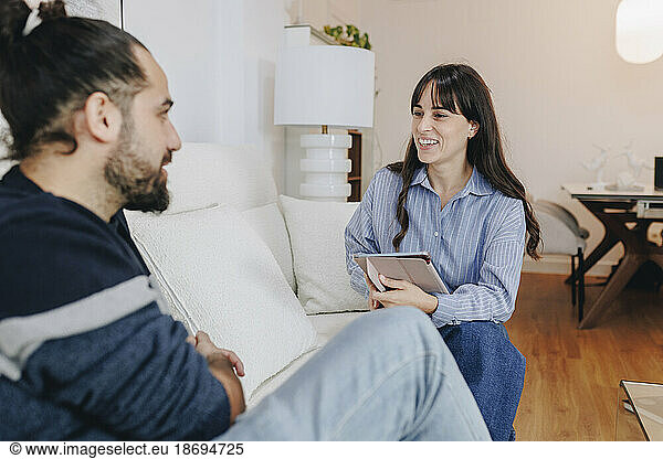 Smiling nutritionist with tablet PC advising client sitting on sofa at home