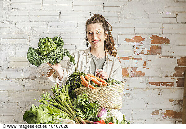 Smiling owner holding kale by vegetables in front of brick wall