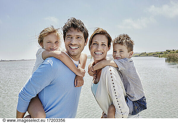 Smiling parents piggybacking children by lake on sunny day