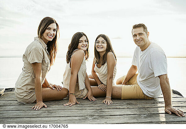 Smiling parents sitting with children on jetty against lake and sky