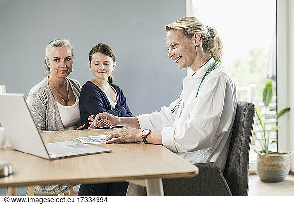 Smiling pediatrician looking at laptop while sitting with girl and woman in office