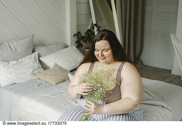 Smiling plus size woman sitting with flowers on bed at home
