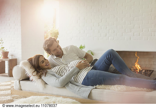 Smiling pregnant couple laying on living room sofa near fireplace