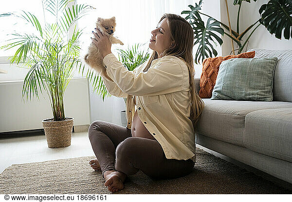 Smiling pregnant woman holding puppy sitting on floor at home