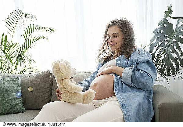 Smiling pregnant woman holding teddy bear sitting on sofa at home