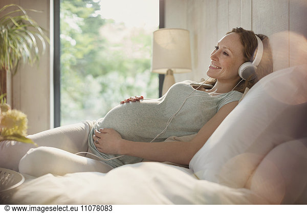 Smiling pregnant woman relaxing listening to music with headphones in bed