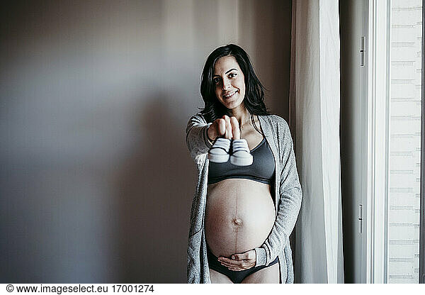 Smiling pregnant woman showing baby booties while standing against wall at home