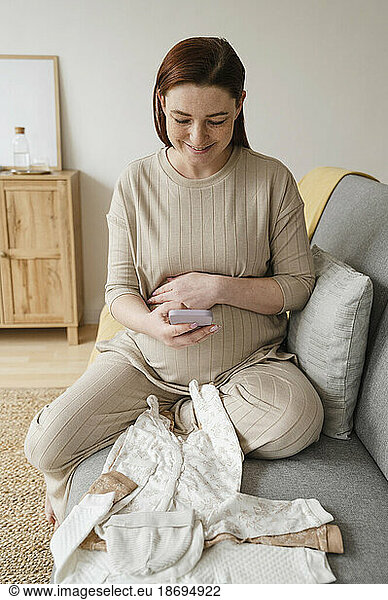 Smiling pregnant woman taking picture of baby clothing sitting on sofa at home