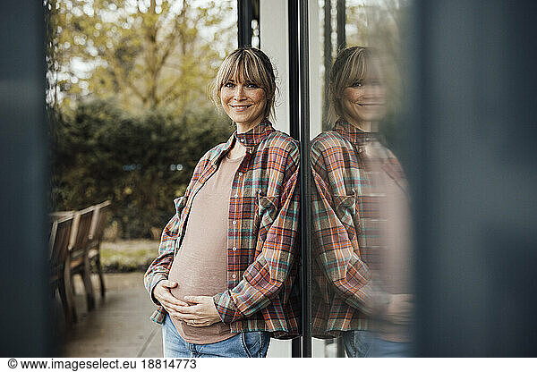 Smiling pregnant woman touching belly leaning on glass door