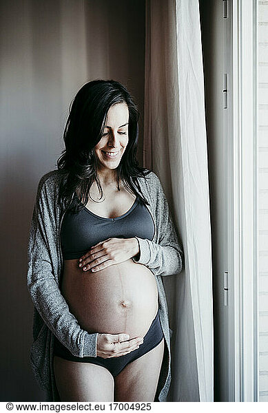Smiling pregnant woman with hands in stomach standing by window at home