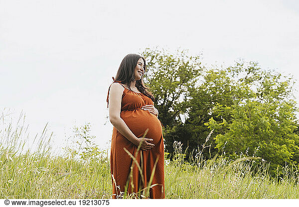 Smiling pregnant woman with hands on stomach standing amidst grass