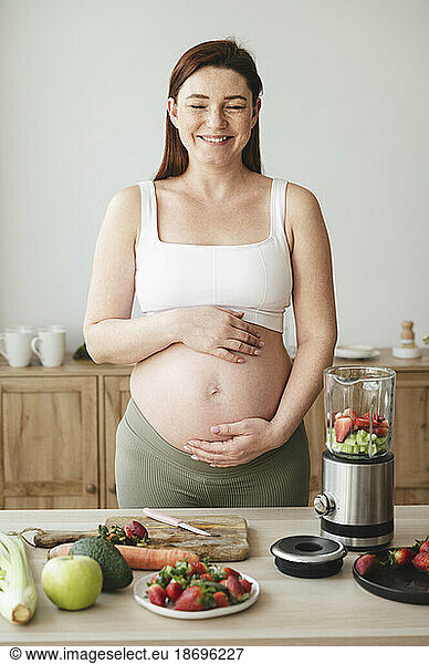 Smiling pregnant woman with hands on stomach standing at home