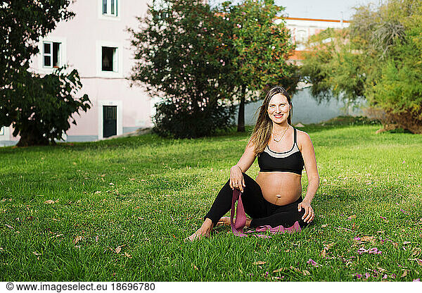 Smiling pregnant woman with resistance band sitting on lawn