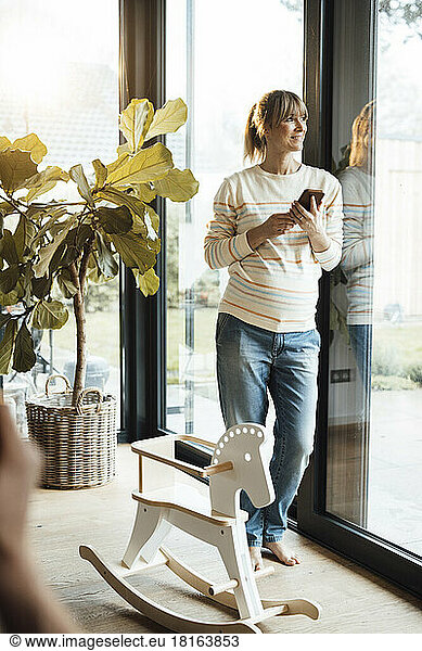 Smiling pregnant woman with smart phone leaning on door at home