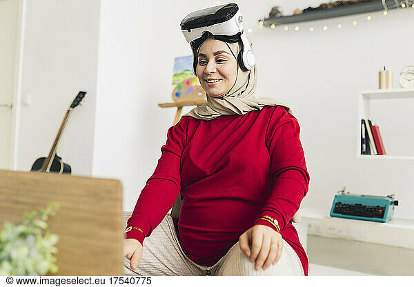 Smiling pregnant woman with VR glasses using laptop in living room