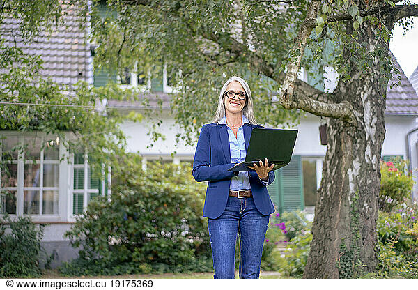 Smiling real estate agent standing with laptop in front of house