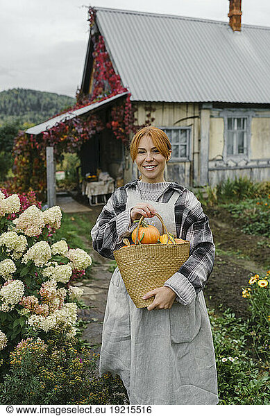 Smiling redhead farmer carrying basket of organic pumpkins at farm