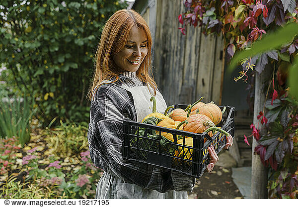 Smiling redhead farmer carrying crate of pumpkins at farm