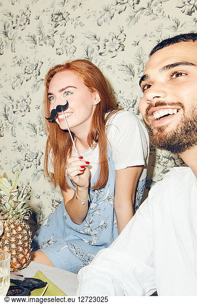 Smiling redhead woman holding mustache prop while sitting by male friend during party at home