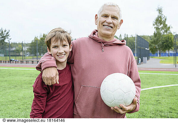 Smiling retired senior man holding soccer ball with arm around grandson