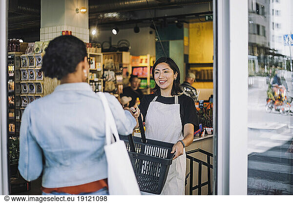 Smiling sales clerk giving shopping basket to female customer arriving at convenience store