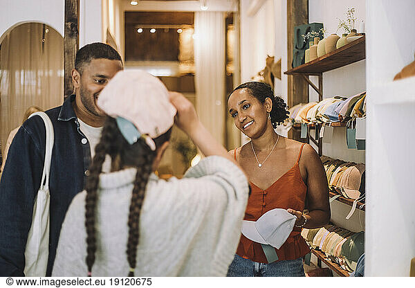 Smiling saleswoman and man looking at girl wearing cap in fashion clothing store